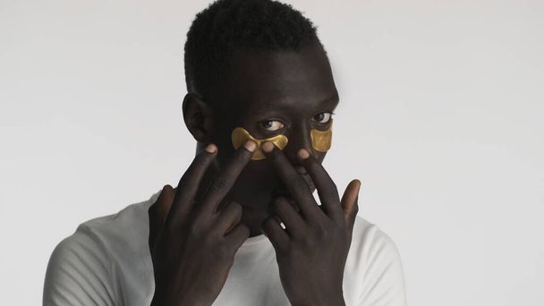 Atractivo chico afroamericano aplicando parches de ojos dorados aislados sobre fondo blanco. Tratamiento facial  - Foto, imagen