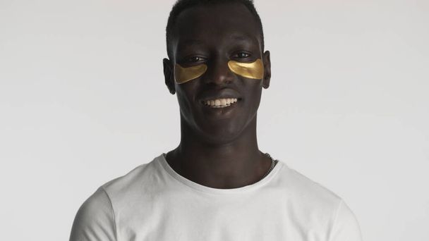 Attractive African American guy with gold eye patches smiling on camera over white background. Facial treatment  - Photo, Image
