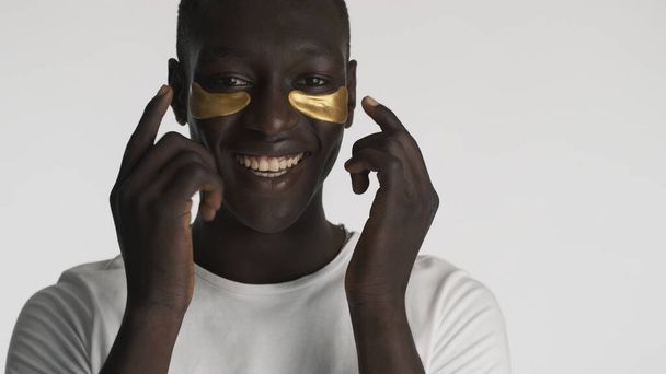 Aantrekkelijke vrolijke Afro-Amerikaanse man met gouden ooglapjes die er blij uitzien over een witte achtergrond. Gezichtsbehandeling  - Foto, afbeelding