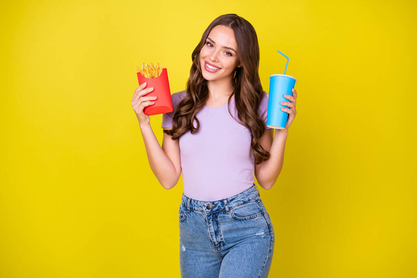 Portrait of pretty cute cheerful wavy-haired girl eating tasty french fries drinking soda isolated on bright yellow color background - 写真・画像