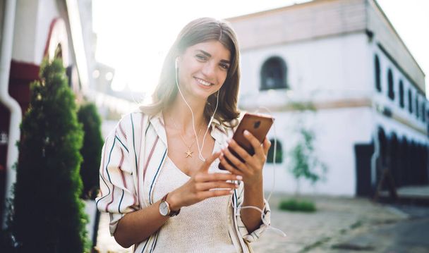 Half length portrait of happy woman in electronic equipment dialing contact number for communicate, cheerful hipster girl in earphones listening positive music podcast from smartphone application - Fotoğraf, Görsel