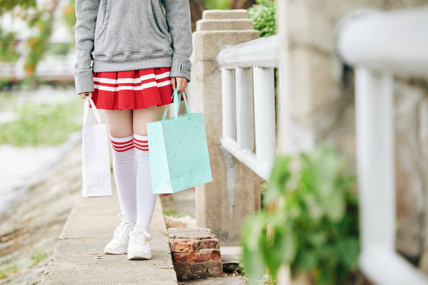 Oříznutý obrázek školačky v krátké sukni a vysoké ponožky drží papírové tašky při chůzi venku - Fotografie, Obrázek