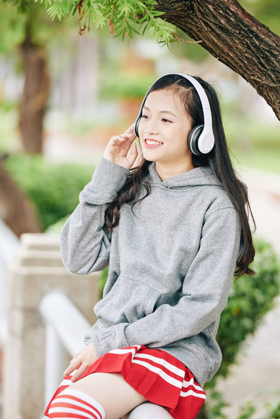 Счастливая симпатичная азиатская студентка, наслаждающаяся хорошей музыкой в наушниках во время отдыха на свежем воздухе - Фото, изображение