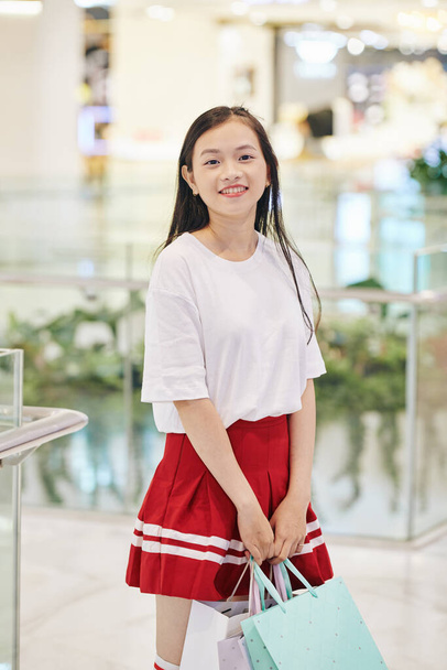 Красивая улыбающаяся девочка-подросток стоит в торговом центре с сумками в руках - Фото, изображение