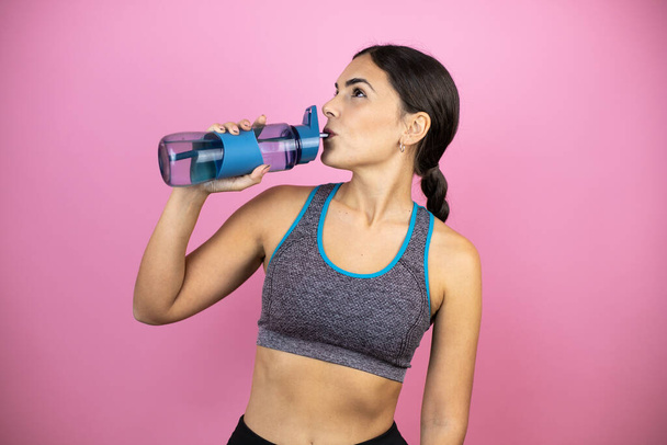 Mladá krásná žena na sobě sportovní oblečení přes izolované růžové pozadí s úsměvem šťastný a přirozený, pití čerstvé vody z láhve. - Fotografie, Obrázek