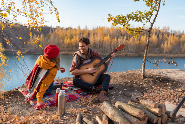 Bir çift, sonbahar ağaçları ve nehrin arka planına karşı doğada dinleniyor. Adam gitar çalıyor. - Fotoğraf, Görsel