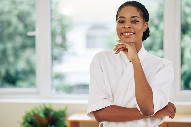 Portrait de jolie jeune femme noire en serviette blanche douce debout dans le salon de spa et souriant à la caméra - Photo, image