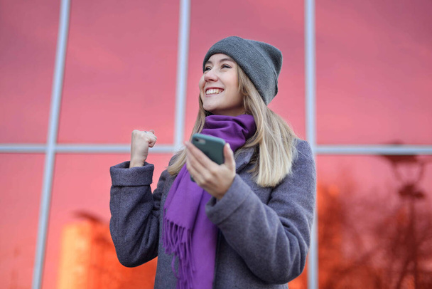 Feliz mujer excitada casual celebrando el éxito sosteniendo el teléfono móvil. se encuentra en la calle calurosamente vestido con un sombrero gris y una bufanda púrpura cerca de un centro comercial sobre un fondo rojo brillante. - Foto, imagen