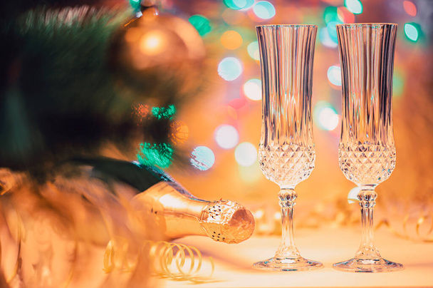 Prázdné křišťálové sklenice a uzavřená láhev šampaňského na pozadí vánočního stromku a barevná světla věnce v mělké hloubce pole. Koncepce nového roku a vánoční prázdniny. - Fotografie, Obrázek