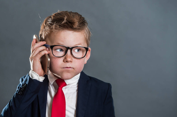 School child boy in business suit and red tie and glasses with cell phone. Funny little businessman. 7 years old kid leader. Leadership, success, creative and innovative business concept - Valokuva, kuva