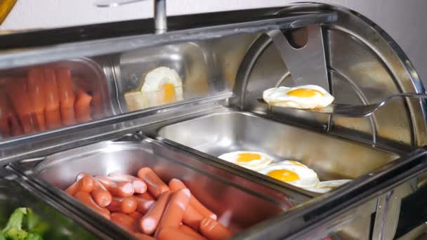 Buffet verwarmde trays met worsten, gekookte broccoli en gebakken eieren in het luxe restaurant van het hotel. Alles inbegrepen. Buffetvoedsel. Ober plaatst stuk gebakken ei. Het hotel serveert een ontbijtbuffet. 4 k video - Video