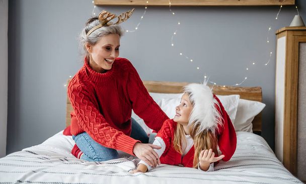 Grand-mère avec petite-fille dans le chapeau rouge de Santa sont allongés sur le lit et riant à Noël. Famille s'amuser pendant les vacances de Noël - Photo, image