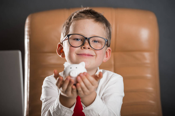 Child calculates savings with pig bank in his hands. Child businessman with earned money in big bag. Ideas how to earn money. Funny little kid boss sitting in leather armchair with laptop - Photo, image