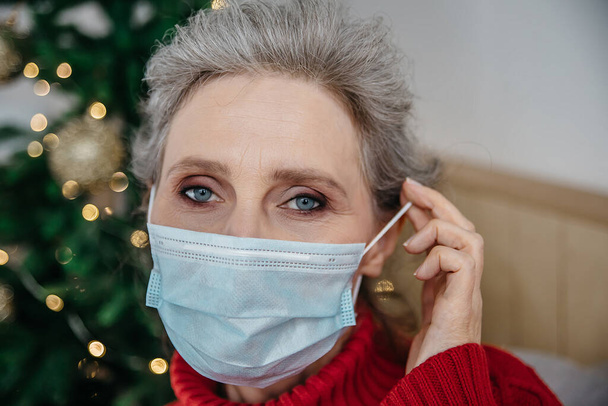Zamknij portret dojrzałej kobiety zakładającej maskę w domu choinkę na tle. Starsza kobieta w masce ochronnej z powodu pandemii covid-19 w Boże Narodzenie. - Zdjęcie, obraz