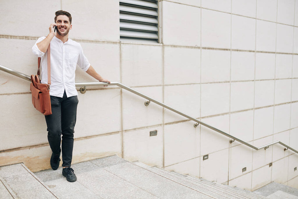 Positive young man standing on steps and talking on phone with friend or coworker - Photo, Image