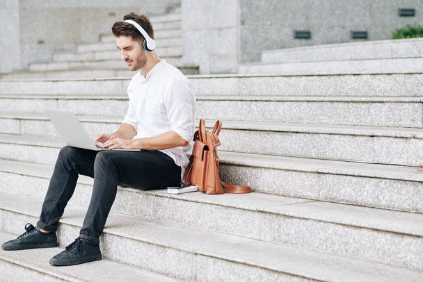 Handsome young programmer in headphones sitting on steps and coding on laptop - Foto, Imagen