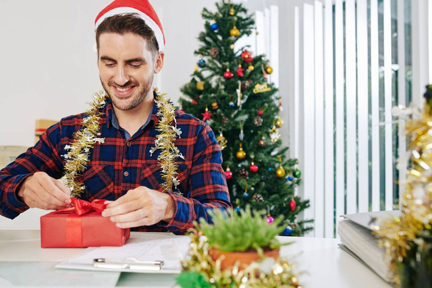彼のオフィスのテーブルに座っているときにクリスマスプレゼントを包む興奮ハンサムなビジネスマン - 写真・画像