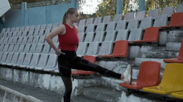 nuori valkoihoinen nainen urheiluvaatteissa venyttelemässä jalkojaan stadionin tribuunissa - Materiaali, video