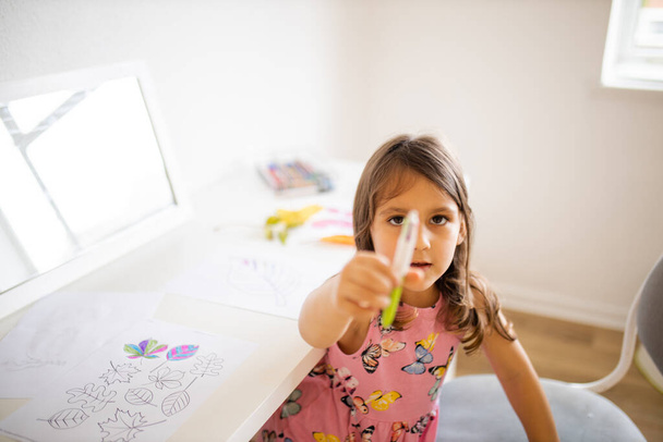 Mała dziewczynka przy stole z kolorowanką liście zdjęcia pokazują długopis - Zdjęcie, obraz