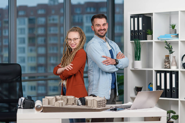 Handsome professional young engineer stands back to back with pretty female colleague with dreadlocks at design bureau. - Foto, Imagen