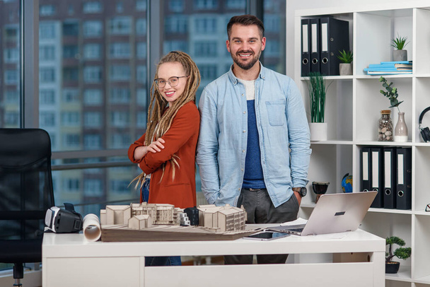 Handsome professional young engineer stands back to back with pretty female colleague with dreadlocks at design bureau. - Foto, Imagem