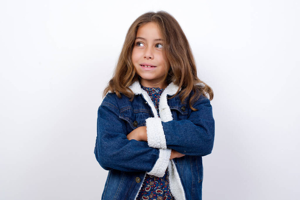 Dreamy rest relaxed Little caucasian girl with beautiful blue eyes wearing denim jacket standing over isolated white background crossing arms, looks good copyspace - Foto, imagen
