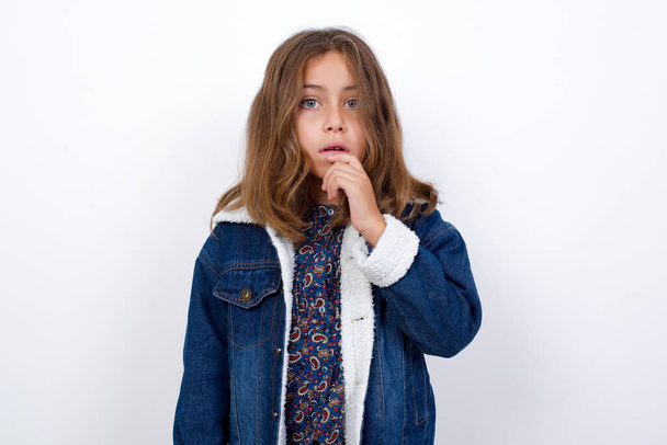 Nervous puzzled Little caucasian girl with beautiful blue eyes wearing denim jacket standing over isolated white background opens mouth from surprise, reacts on sudden news. - Photo, Image