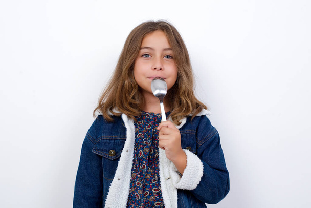 Πολύ πεινασμένο κοριτσάκι Καυκάσιος με όμορφα μπλε μάτια φορώντας τζιν σακάκι στέκεται πάνω από απομονωμένο λευκό φόντο κρατώντας κουτάλι στο στόμα όνειρο νόστιμο γεύμα - Φωτογραφία, εικόνα