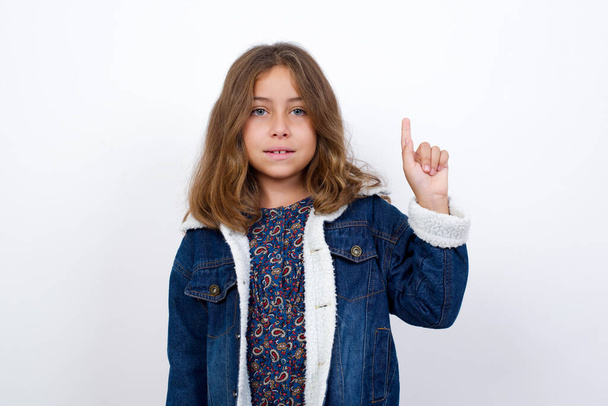 Little caucasian girl with beautiful blue eyes wearing denim jacket standing over isolated white background showing and pointing up with finger number one while smiling confident and happy. - Photo, Image