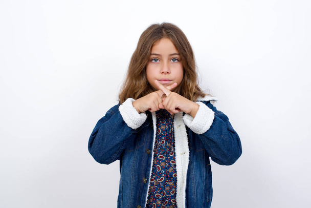 Маленькая кавказская девочка с красивыми голубыми глазами в джинсовой куртке, стоящей на изолированном белом фоне, отказалась от злого выражения, скрестив пальцы. - Фото, изображение