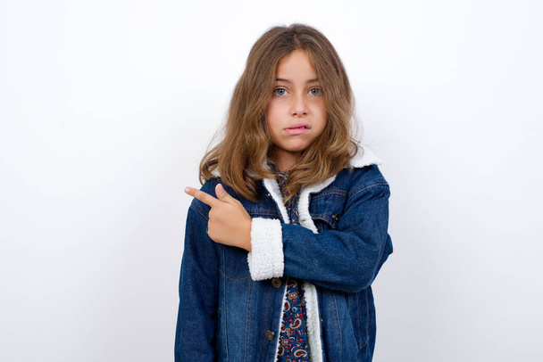 Маленька біла дівчинка з красивими блакитними очима в джинсовій куртці, що стоїть над ізольованим білим тлом. Вказуючи на занепокоєння і нервовий з переслідувачем, занепокоєння і несподівану концепцію
. - Фото, зображення