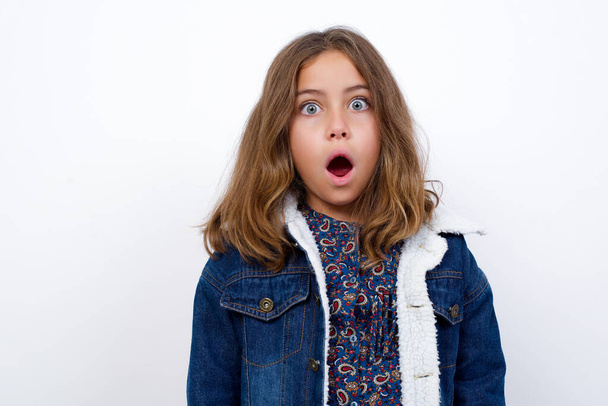 Oh my God. Surprised  Little caucasian girl with beautiful blue eyes wearing denim jacket standing over isolated white background stares at camera with shocked expression exclaims with unexpectedness, - Foto, imagen