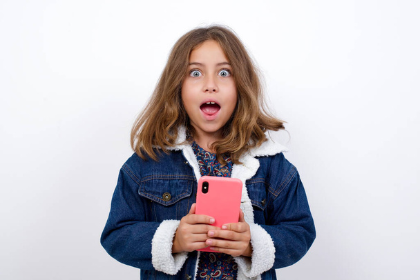 Copyspace photo of cheerful nice cute charming Little caucasian girl with beautiful blue eyes wearing denim jacket standing over isolated white background holding phone in his hands while - Фото, зображення