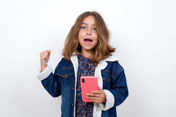 Portrait of Little caucasian girl with beautiful blue eyes wearing denim jacket standing over isolated white background holding in hands cell celebrating  - Photo, Image