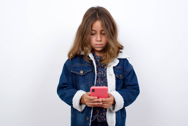 Porträt eines aufgeregten kleinen kaukasischen Mädchens mit schönen blauen Augen, das Jeansjacke trägt, vor isoliertem weißem Hintergrund steht und augenzwinkernd Smartphone benutzt, lesen Sie Nachrichten aus dem sozialen Netzwerk  - Foto, Bild