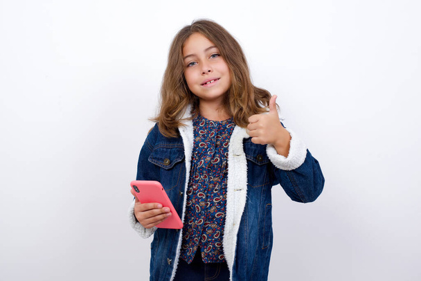 Portrait of Little caucasian girl with beautiful blue eyes wearing denim jacket standing over isolated white background using and texting with smartphone  happy with big smile doing ok sign, thumb up with fingers, excellent sign - Photo, Image