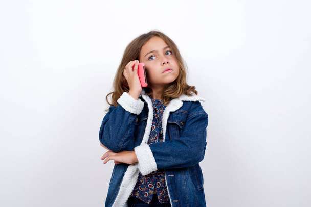 Sad Little caucasian girl with beautiful blue eyes wearing denim jacket standing over isolated white background talking on smartphone. Communication concept. - Zdjęcie, obraz