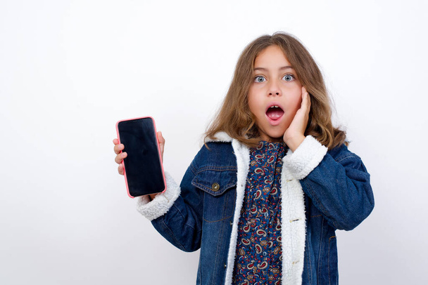 Little caucasian girl with beautiful blue eyes wearing denim jacket standing over isolated white background hold hand modern technology use touch face palm astonished impressed scream wow omg unbelievable unexpected - Foto, Imagen