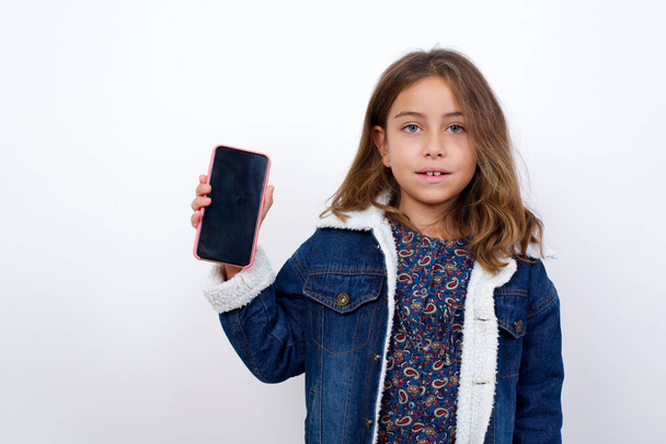 Улыбающаяся маленькая кавказская девочка с красивыми голубыми глазами в джинсовой куртке, стоящей на изолированном белом фоне. Держите мобильный телефон с пустым экраном - Фото, изображение