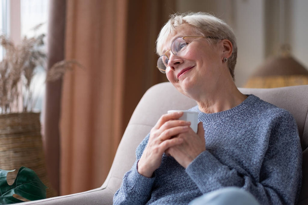 Старшая белая женщина расслабляется, выпивая чашку чая в кресле дома - Фото, изображение