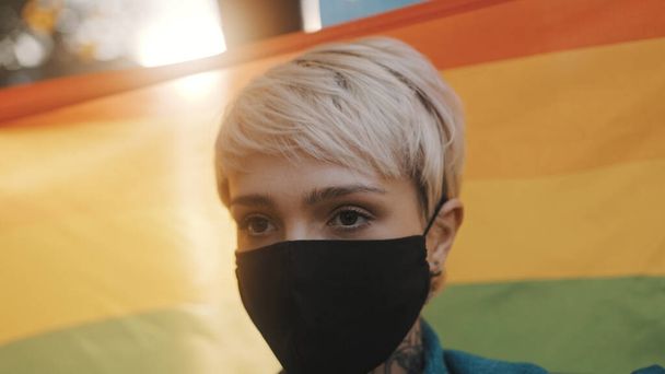 Proud young blond woman with face mask waving rainbow flag. Close up - Photo, Image