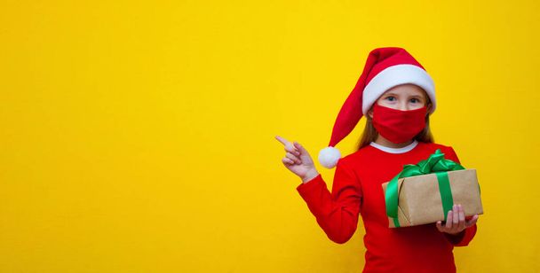 Dziewczyna w kapeluszu Mikołaja, w czerwonym T-shircie, w masce medycznej w rękach trzyma prezent na Boże Narodzenie i Nowy Rok i wskazuje palcem na miejsce na reklamę.  - Zdjęcie, obraz