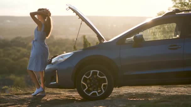 Молодая женщина-водитель стоит рядом со сломанной машиной с открытым капотом, осматривая двигатель и ожидая помощи. - Кадры, видео