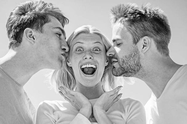 Blonde happy girl getting kisses from two handsome boys. Charming woman standing between two friends. Best friends concept. Group of friends at clear sky background. - Photo, image