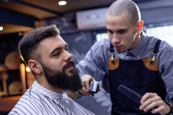 Estilo de barba e corte com aparador. Homem jovem hipster barbudo recebendo corte de cabelo / barba cortada por cabeleireiro profissional em uma barbearia moderna. Conceito de publicidade de barbearia. Movembro. - Foto, Imagem