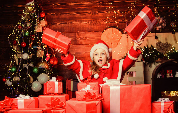Child happy excited girl find gifts near christmas tree. Merry christmas. Happy childhood concept. Kid wear santa hat hold wrapped christmas gift box. Santa brought me gifts. Happiness and joy - Valokuva, kuva