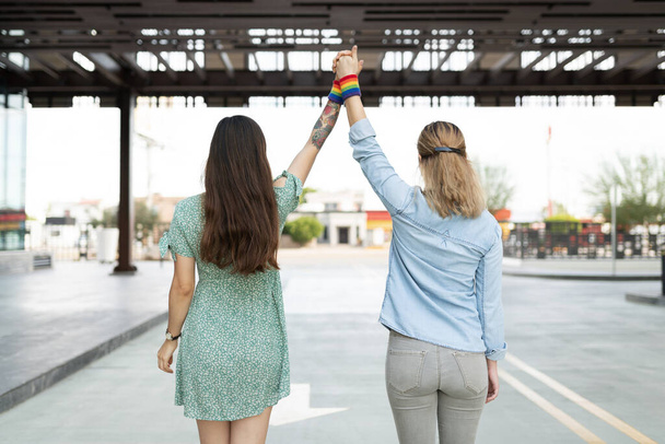 Multiracial lesbiana pareja desde detrás levantando sus manos juntos y usando un arco iris gay brazo banda  - Foto, imagen