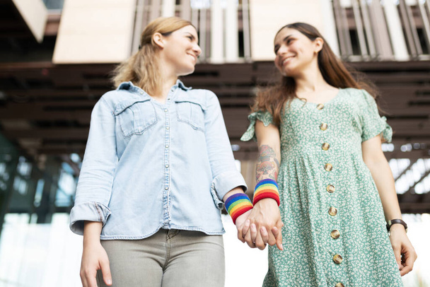 Low angle of a caucasian gay woman and a latin lesbian woman standing and holding hands with rainbow colored wristbands - Photo, Image