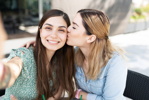 Blondýny roztomilý gayové mladá žena líbání na tvář její lesbičky latinsky přítelkyně zatímco ona bere selfie - Fotografie, Obrázek