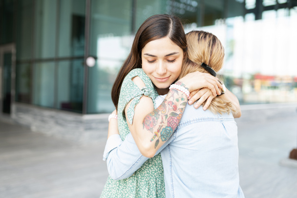 Latein junge schwule Frau sieht friedlich und glücklich aus, während sie ihre blonde lesbische Freundin außerhalb eines Gebäudes umarmt - Foto, Bild